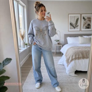 A woman in a bedroom takes a mirror selfie, wearing a light gray crewneck sweatshirt with a gold celestial moth design and light wash jeans.