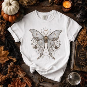 A white short-sleeved t-shirt with a gray and gold celestial moth design is displayed flat on a dark wooden surface with autumn decor.