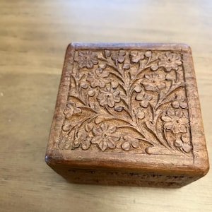 May include: A square wooden box featuring a detailed hand-carved floral pattern on the lid. The natural brown wood grain is visible across the surface, and the carving depicts a cluster of flowers with stems and leaves covering the top panel.