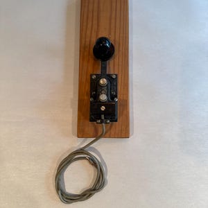 May include: Vintage telegraph key mounted on a rectangular wooden base. The key features a black knob and a metal mechanism. A gray, braided wire extends from the base. The wood has a natural grain.