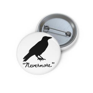 May include: A round pin-back button featuring a white background with a black silhouette of a raven. Below the bird, the word "Nevermore." is printed in a black cursive script. The back of the button is silver metal with a pin mechanism.