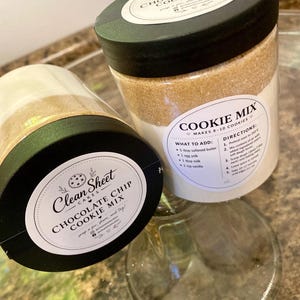 Könnte beinhalten: Zwei transparente Gläser mit Clean Sheet Cakes Schokoladen-Chip-Keksmischung. Die Gläser haben schwarze Deckel und weiße Etiketten mit Text. Ein Glas steht aufrecht, das andere ist geneigt. Die Gläser sind mit einer Mischung aus Zutaten gefüllt.