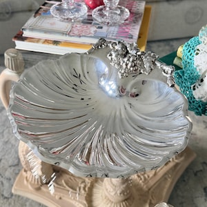 May include: A silver-toned metal dish shaped like a scallop shell, featuring a decorative relief of a grape cluster and leaves at the top edge. The surface of the dish has a polished, reflective finish with deep, radiating fluted grooves.