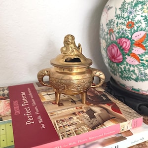 May include: A small, antique brass incense burner with a lid and three legs. The burner has elephant-shaped handles and decorative carvings. It sits atop a stack of books with the title "Perfect Patterns" and a floral vase in the background.
