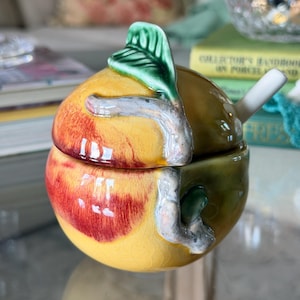 May include: A ceramic lidded jar shaped like a peach with a glossy yellow, orange, and red finish. The lid features a sculpted green leaf and a branch detail that extends onto the base. A small white ceramic spoon rests inside the jar.