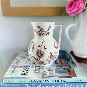 May include: A cream-colored ceramic pitcher featuring a dark red floral and geometric transferware pattern. The pitcher has a rounded body, a flared rim, and a single angular handle. It is decorated with detailed botanical motifs and small vignettes.