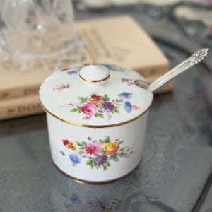 May include: A white porcelain sugar bowl with a matching lid and a silver-toned metal spoon. The bowl and lid feature colorful floral patterns of roses, pansies, and daisies, accented with gold trim along the rims and the lid handle.
