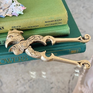 May include: A brass nutcracker shaped like a bird with detailed feather engravings on the handles. The tool features a hinged mechanism with a beak-like head and curved, scroll-ended handles, showcasing a metallic gold finish and a textured surface.