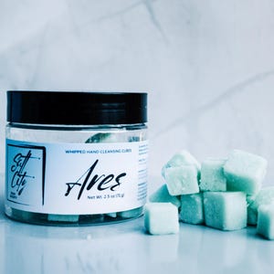 May include: A clear plastic jar with a black lid containing light green, cube-shaped hand cleansing soap. The white label features the text Salt City, Ares, and Whipped Hand Cleansing Cubes, with a net weight of 2.5 oz (71 g) printed on the front.