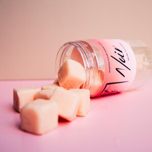 May include: A clear plastic jar lying on its side, spilling out several pale peach-colored, cube-shaped sugar scrub pieces. The jar features a pink label with the text "Noir" and "Whipped Sugar Scrub Cubes" printed in black, along with "Net Wt. 2.5 oz."