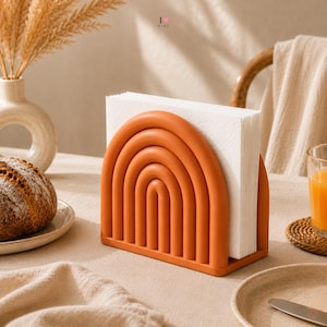 May include: A terracotta-colored napkin holder featuring a series of concentric arched bars that form a rainbow shape. The holder is filled with a stack of white square paper napkins, standing upright on a flat base with a smooth matte finish.