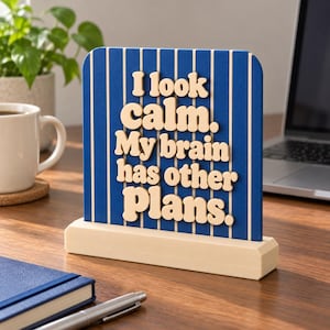 May include: A rectangular desk sign with a blue background featuring vertical white pinstripes. The sign displays raised, light-colored, rounded lettering that reads: I look calm. My brain has other plans. It sits on a light wood base.