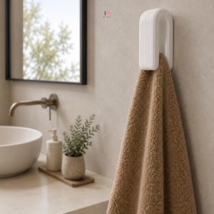 Op de afbeelding: Een witte, U-vormige wandhaak met een geribbelde textuur, gemonteerd aan een muur en voorzien van een gevouwen, getextureerde bruine handdoek. Boven de haak staat de kleine tekst I heart MIMO in zwart en rood, waarbij een hartje de letter O vervangt.