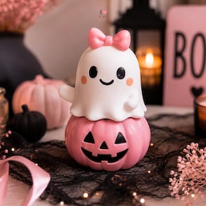 May include: A white ghost figurine with a pink bow on its head, perched atop a pink jack-o'-lantern pumpkin. The ghost has black eyes, a smiling mouth, and small pink cheeks. The pumpkin features a carved black face with triangular eyes and a grin.