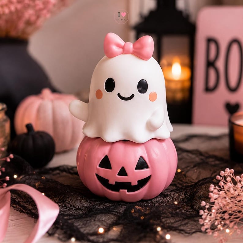 May include: A white ghost figurine with a pink bow on its head, perched atop a pink jack-o'-lantern pumpkin. The ghost has black eyes, a smiling mouth, and small pink cheeks. The pumpkin features a carved black face with triangular eyes and a grin.