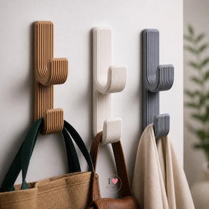 May include: Three vertical wall hooks in tan, white, and dark gray, each featuring a ribbed texture and two upward-curving prongs. The tan hook holds a brown fabric bag, the white hook holds a brown leather bag, and the gray hook holds a beige cloth.