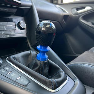 Könnte beinhalten: Ein Schaltknauf aus Kohlefaser mit blauem Sockel ist in einem Auto eingebaut. Die Schalthebelmanschette ist aus schwarzem Leder mit blauen Nähten. Der Innenraum des Autos ist schwarz mit silbernen Akzenten.
