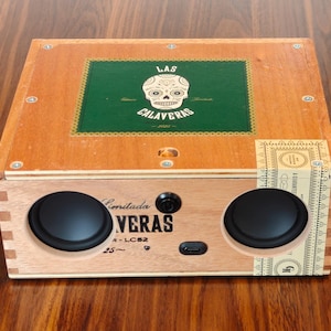 May include: A rectangular wooden cigar box repurposed as a speaker, featuring two circular black speakers on the front. The top lid displays a green square label with a white skull illustration and the text LAS CALAVERAS 2022 in white lettering.
