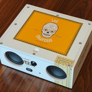 May include: A white rectangular speaker box featuring a yellow top label with a black and white sugar skull illustration and the text Las Calaveras. The front panel includes two circular black speaker drivers, a central power button, and a port.
