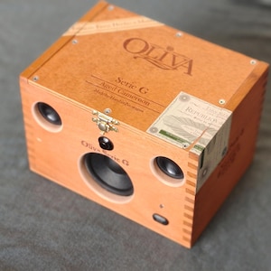 May include: A custom-built speaker system housed in a wooden Oliva Serie G cigar box. The box is light brown with black speaker components and a brass clasp. The box has the text "Oliva Serie G Aged Cameroon" on the front.