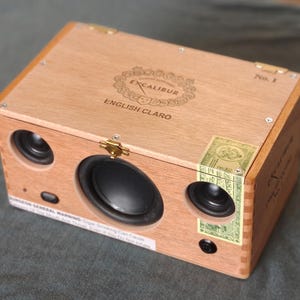 May include: A handmade wooden speaker crafted from a cigar box. The box features the text "EXCALIBUR ENGLISH CLARO" and has two small speakers and one larger speaker. The speaker has gold-colored corner protectors and a gold clasp.