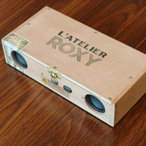 May include: A rectangular wooden box speaker featuring the text L'ATELIER ROXY printed on the top surface. The device includes two circular black speaker drivers on the front panel, a gold-colored metal latch, and a small black charging port.