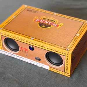 May include: A rectangular speaker system housed in a wooden cigar box. The box is brown with yellow trim and features the "PUNCH" logo. Two black speakers are visible on the front, along with a blue light and a charging port. The box has text that reads "SURGEON GENERAL WARNING".