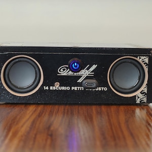 May include: A black rectangular portable speaker featuring two circular speakers, a central blue illuminated power button, and a USB-C charging port. The front panel displays the white text Davidoff and 14 ESCURIO PETIT ROBUSTO.