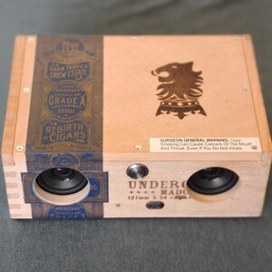 May include: A wooden box speaker with a cigar box design. The box features a blue and gold label with text and a decorative emblem. Two speakers are visible on the front, along with a control knob and a warning label.