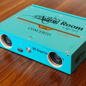 May include: A rectangular turquoise box repurposed as a speaker, featuring the text Aging Room by Rafael Nodal, Concerto, and Quattro. The front panel includes two circular black speaker drivers, a small charging port, and a metal power button.