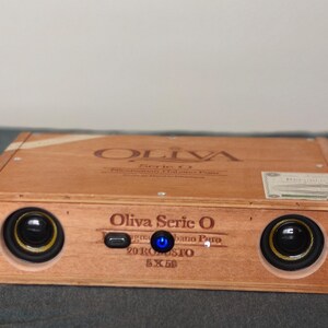 May include: A handmade speaker system housed in a wooden Oliva Serie O cigar box. The box features two circular speakers with gold trim, a blue power light, and the text "Oliva Serie O" on the front. The wooden box is light brown.