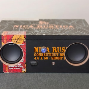 May include: A black speaker box with two circular speakers and the text "NICA RUSTICA CONNECTICUT BR 4.5 X 50 - SHORT M". The box has a decorative top and a colorful label with text. The speakers have a light wood-colored trim.