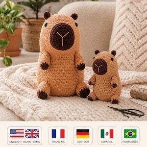 May include: Two handmade crochet capybara plush toys in light tan yarn with dark brown snouts, ears, and feet. The larger capybara stands next to a smaller version, both featuring black bead eyes and a stitched Y-shaped nose detail on their faces.