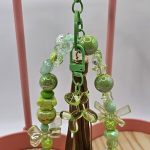 Puede incluir: Un llavero verde con cuentas verdes, un cierre verde y una borla marr&oacute;n. El llavero presenta varias cuentas verdes, incluyendo cuentas redondas, en forma de lazo y de animales, junto con cuentas transparentes e iridiscentes. Se muestra en un soporte rosa.