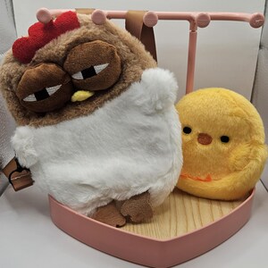 Puede incluir: Una mochila con forma de pollo de peluche con cresta roja, ojos marrones y vientre blanco, junto a un peluche de pollito amarillo. Los art&iacute;culos descansan sobre una bandeja rosa en forma de coraz&oacute;n con base de madera y un expositor de joyas rosa.