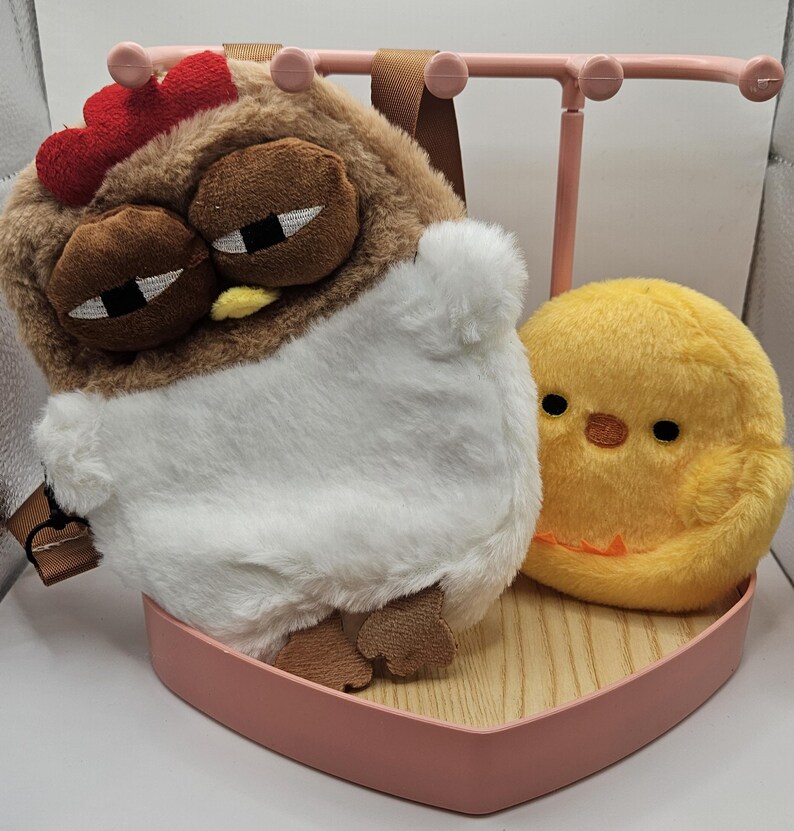 Puede incluir: Una mochila con forma de pollo de peluche con cresta roja, ojos marrones y vientre blanco, junto a un peluche de pollito amarillo. Los art&iacute;culos descansan sobre una bandeja rosa en forma de coraz&oacute;n con base de madera y un expositor de joyas rosa.