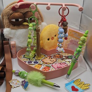 Puede incluir: Una colecci&oacute;n de accesorios y art&iacute;culos de papeler&iacute;a caprichosos. Incluye peluches, llaveros con cuentas, bol&iacute;grafos decorativos y pines esmaltados. Una bandeja rosa en forma de coraz&oacute;n contiene pegatinas con dise&ntilde;os de comida pixelados.