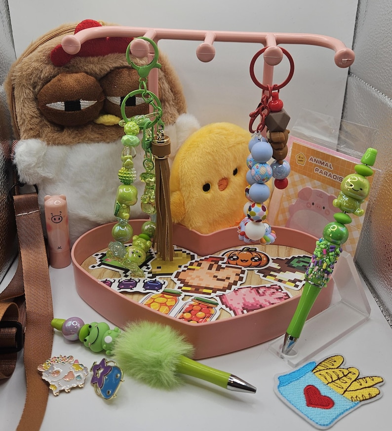 Puede incluir: Una colecci&oacute;n de accesorios y art&iacute;culos de papeler&iacute;a caprichosos. Incluye peluches, llaveros con cuentas, bol&iacute;grafos decorativos y pines esmaltados. Una bandeja rosa en forma de coraz&oacute;n contiene pegatinas con dise&ntilde;os de comida pixelados.