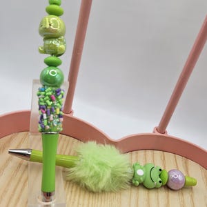 Puede incluir: Una colecci&oacute;n de bol&iacute;grafos decorativos con dise&ntilde;os &uacute;nicos. Un bol&iacute;grafo presenta un personaje verde con forma de rana, otro tiene un acento verde esponjoso y un tercero tiene un dise&ntilde;o verde apilado con chispas de colores. Los bol&iacute;grafos se exhiben en una bandeja de madera.