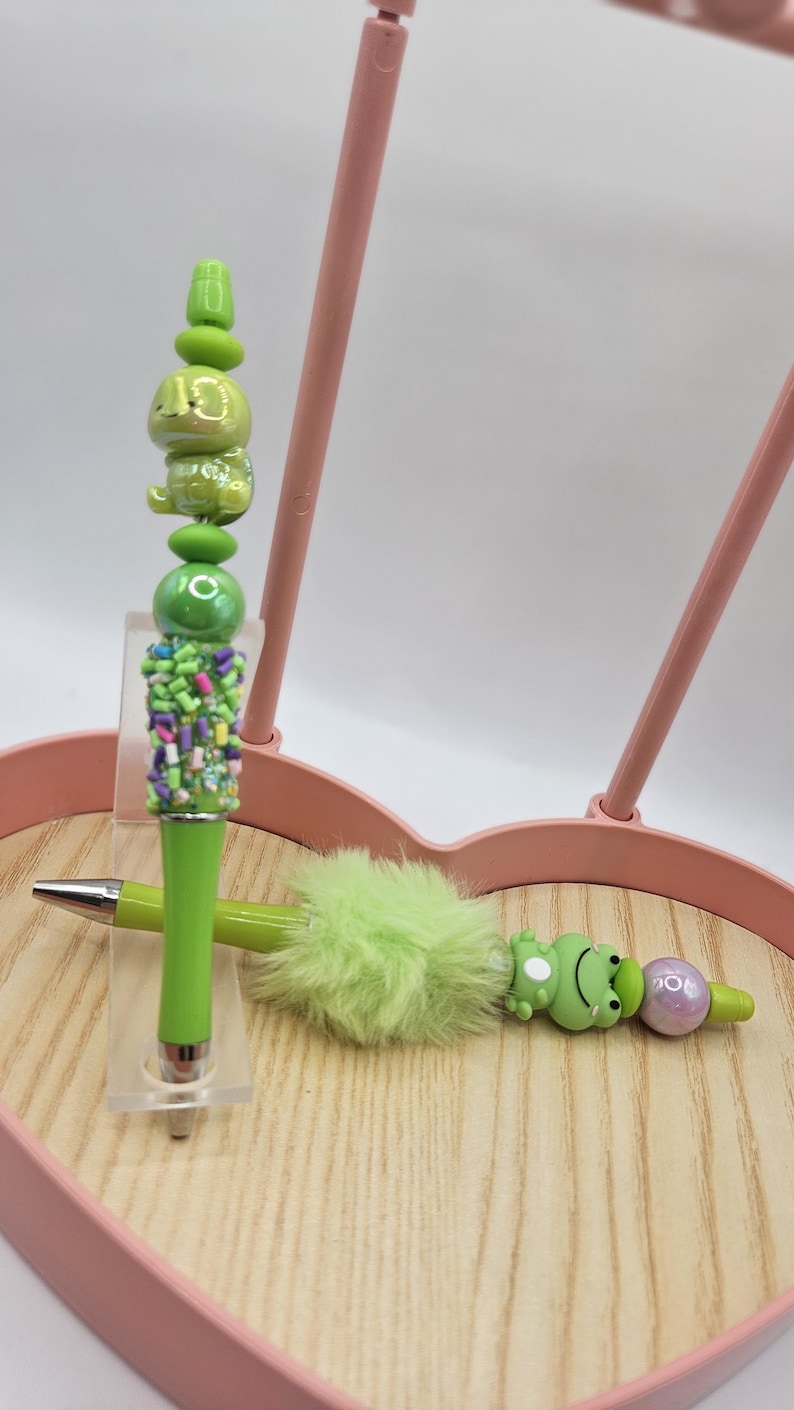 Puede incluir: Una colecci&oacute;n de bol&iacute;grafos decorativos con dise&ntilde;os &uacute;nicos. Un bol&iacute;grafo presenta un personaje verde con forma de rana, otro tiene un acento verde esponjoso y un tercero tiene un dise&ntilde;o verde apilado con chispas de colores. Los bol&iacute;grafos se exhiben en una bandeja de madera.