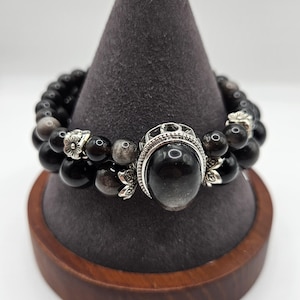 May include: Two black beaded bracelets with silver accents. One bracelet features a large, round, black stone with a silver setting. The other bracelet has smaller black beads and silver floral accents. The bracelets are displayed on a gray cone-shaped stand.