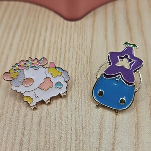 Puede incluir: Dos pines de esmalte sobre una superficie de madera. Un pin es una oveja blanca con acentos coloridos y una corona floral. El otro es un personaje azul y morado con una cabeza en forma de estrella y detalles dorados. Los pines son probablemente para ropa o accesorios.