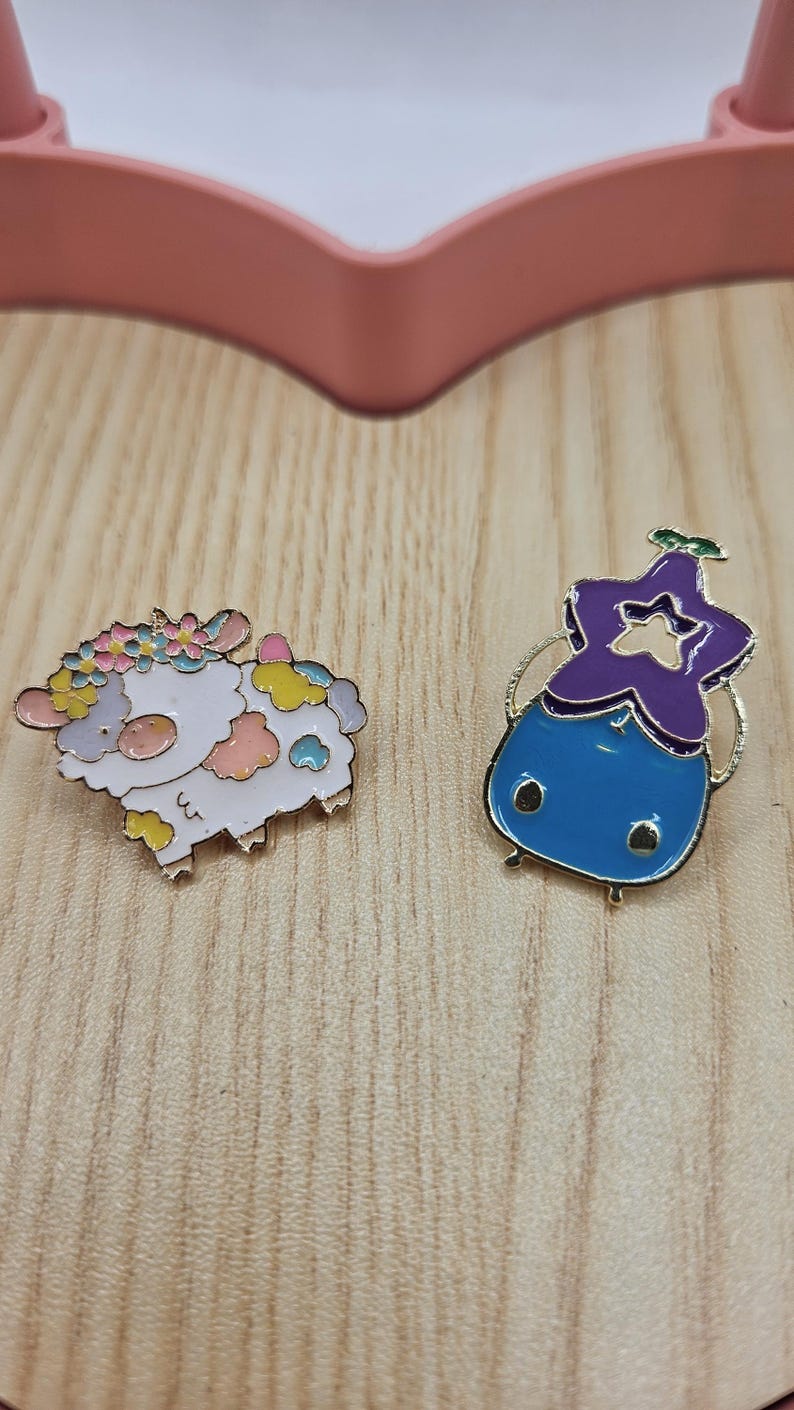 Puede incluir: Dos pines de esmalte sobre una superficie de madera. Un pin es una oveja blanca con acentos coloridos y una corona floral. El otro es un personaje azul y morado con una cabeza en forma de estrella y detalles dorados. Los pines son probablemente para ropa o accesorios.