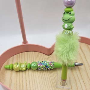 Puede incluir: Un bol&iacute;grafo verde con un dise&ntilde;o decorativo que presenta una rana, cuentas y un acento verde esponjoso. El bol&iacute;grafo se muestra en una bandeja de madera con un marco rosa en forma de coraz&oacute;n. El bol&iacute;grafo mide unos 15 cm de largo.
