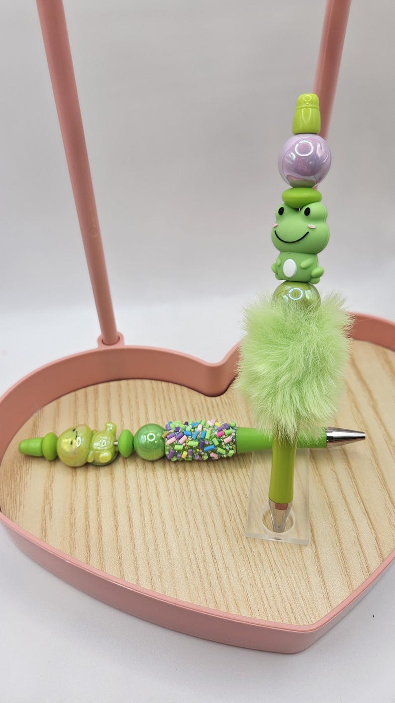 Puede incluir: Un bol&iacute;grafo verde con un dise&ntilde;o decorativo que presenta una rana, cuentas y un acento verde esponjoso. El bol&iacute;grafo se muestra en una bandeja de madera con un marco rosa en forma de coraz&oacute;n. El bol&iacute;grafo mide unos 15 cm de largo.