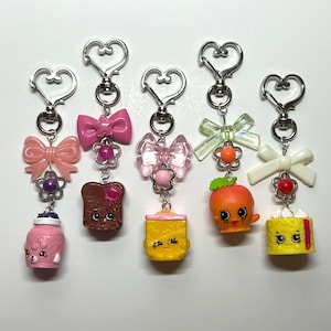 Könnte beinhalten: Fünf Schlüsselanhänger mit Miniatur-Lebensmittelfiguren mit Cartoon-Gesichtern, darunter ein rosa Glas, brauner Toast, ein gelber Block, eine orange Frucht und ein gelber Kuchen. Jeder Anhänger hat einen silbernen herzförmigen Verschluss.