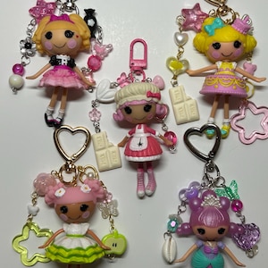 May include: A collection of colorful doll keychains. Each doll has a unique design, with varying hairstyles, outfits, and accessories like stars, hearts, and candy. The keychains have metal clasps for easy attachment.
