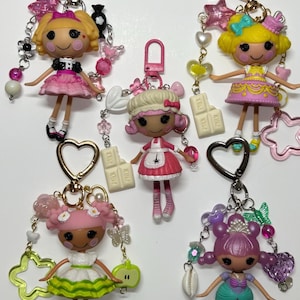 May include: Five Lalaloopsy doll keychains with heart-shaped clasps. Each doll features unique outfits and hair colours, including pink, yellow, and purple. The keychains are decorated with various charms like stars, hearts, butterflies, and miniature chocolate bars.