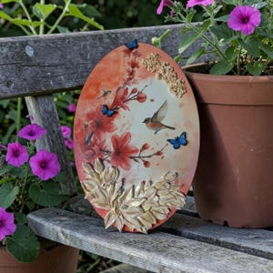 Könnte beinhalten: Eine ovale Wandplakette mit einer gemalten Szene eines kleinen braunen Vogels im Flug zwischen roten Blumen. Die Plakette ist mit drei kleinen blauen Schmetterlingsakzenten und einer Ansammlung goldfarbener Blätter unten und oben verziert.