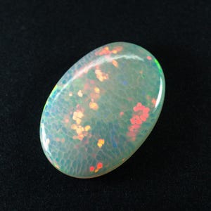 Op de afbeelding: Een ovaalvormige opaal edelsteen met een lichtgroene basiskleur. De steen heeft een textuur met kleine, kleurrijke vlekken van oranje, rood en geel. De opaal is geplaatst tegen een zwarte achtergrond.